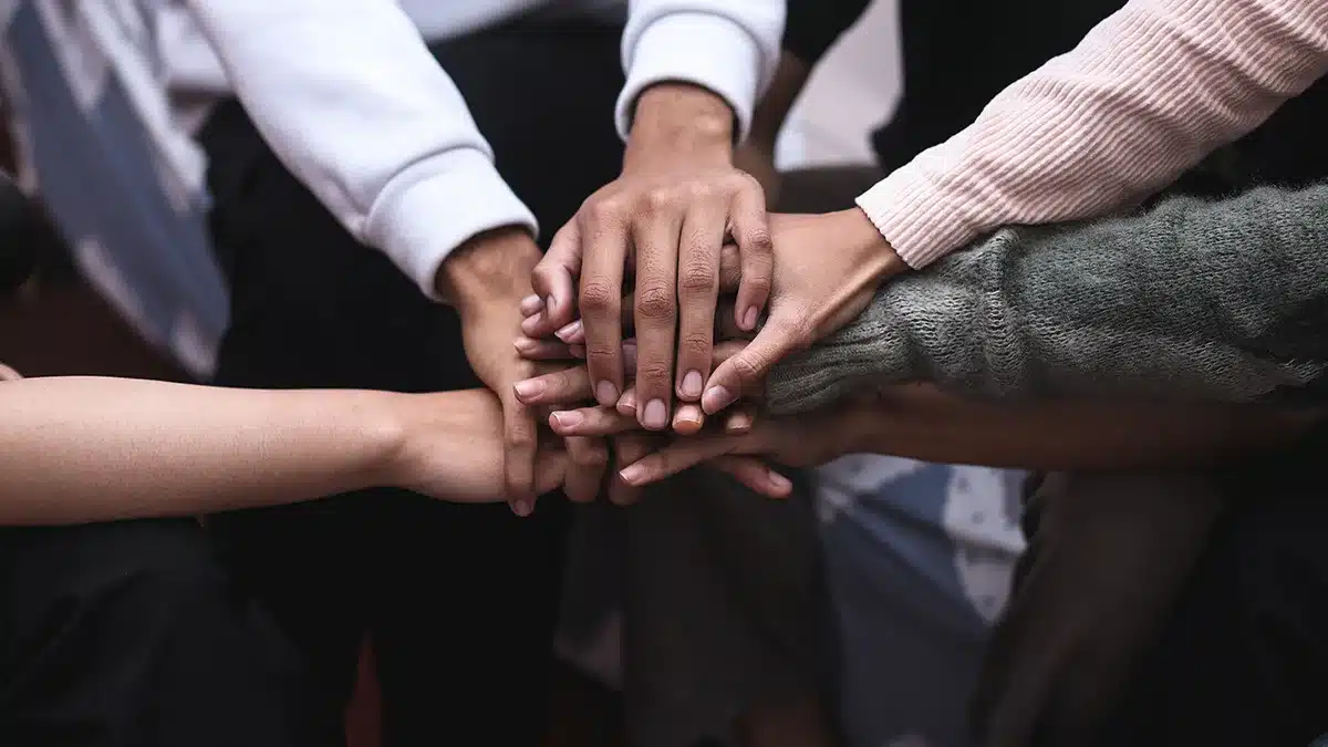 Multiple hands stacked on top of each other in a show of unity and support. The image focuses on the hands, with some individuals wearing long sleeves and others in different types of clothing, indicating a diverse group coming together.