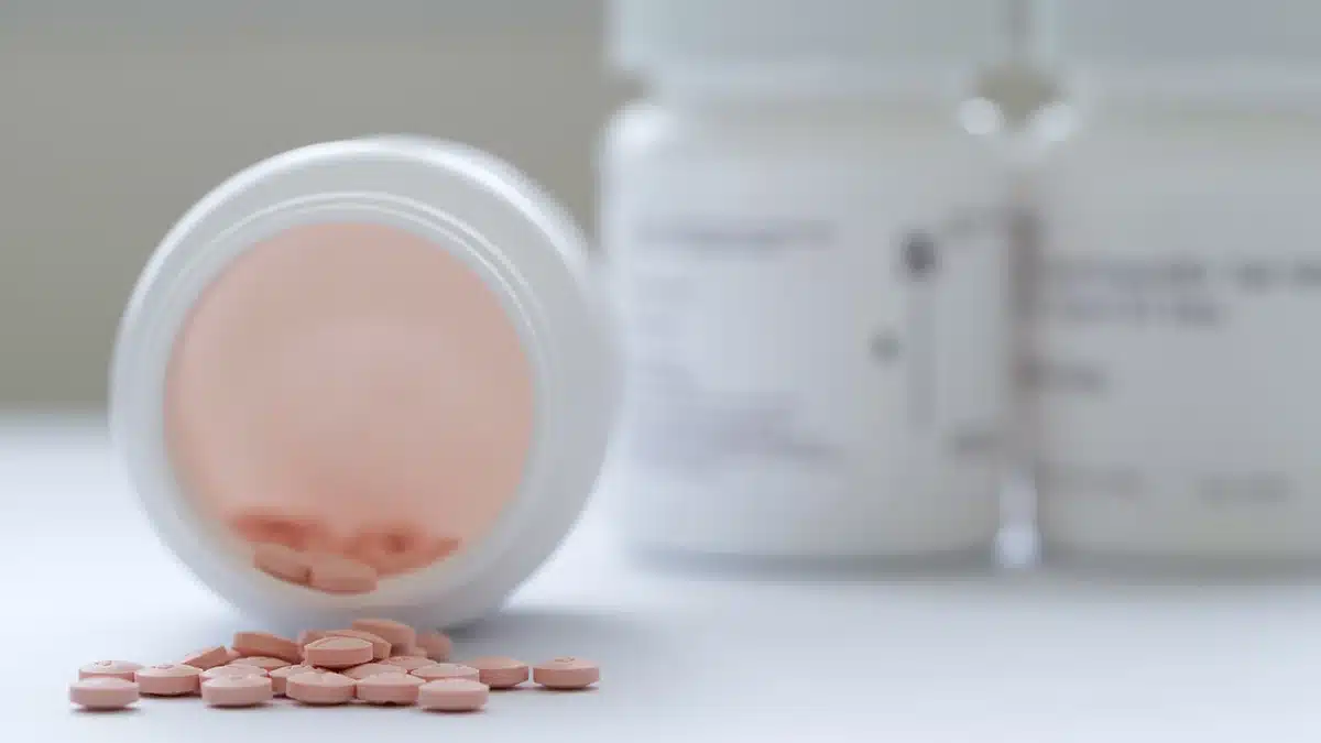 A close-up of a prescription pill bottle tipped over, with pink tablets spilling out, alongside blurred pill bottles in the background