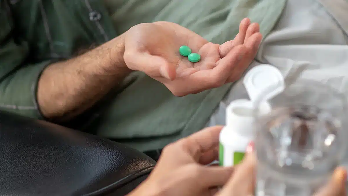 A close-up shot shows a person holding two green pills in one hand, while the other hand holds a bottle of medication with the cap off.