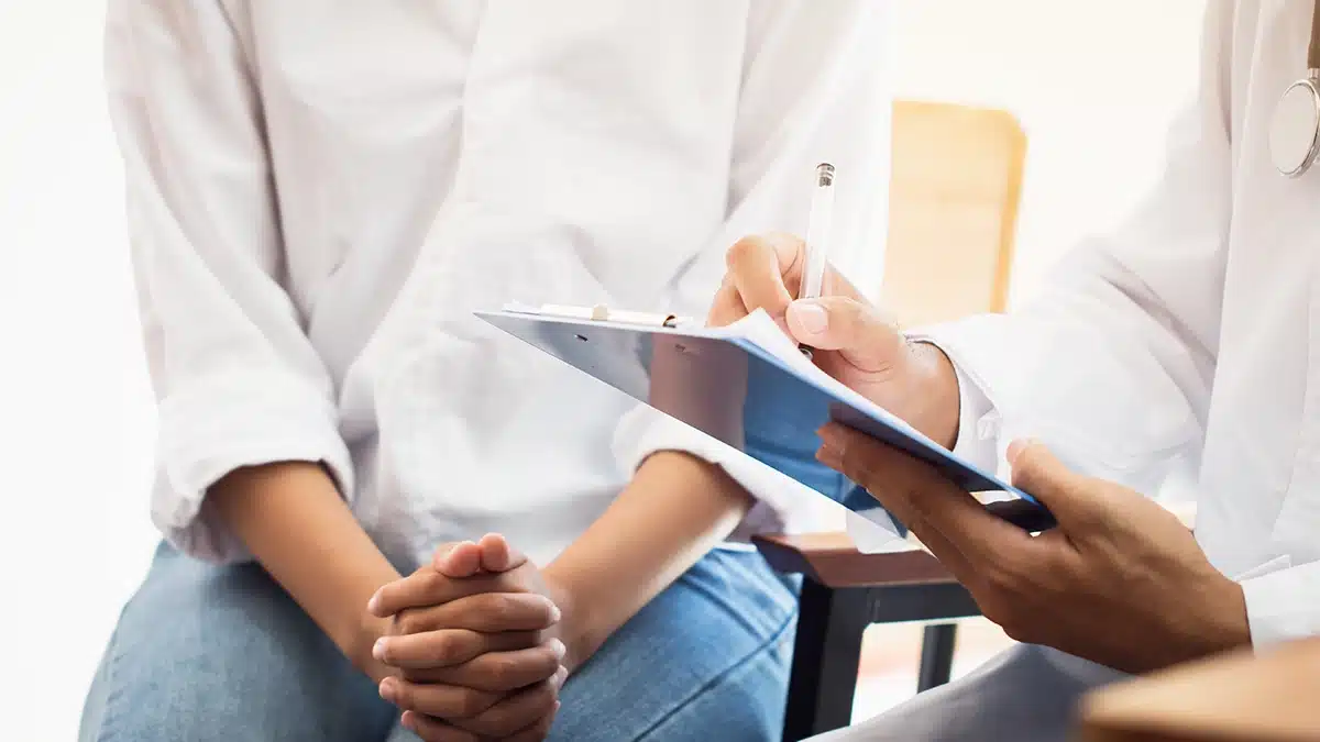 A close-up of a doctor taking notes on a clipboard while talking to a patient, focusing on the hands of both individuals during the consultation