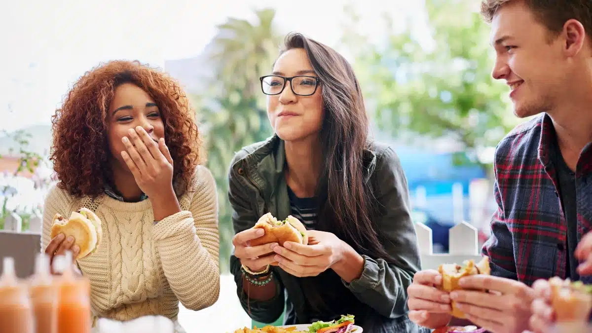 A group of three friends enjoying a meal together outdoors, laughing and talking while holding sandwiches, creating a cheerful and lively atmosphere