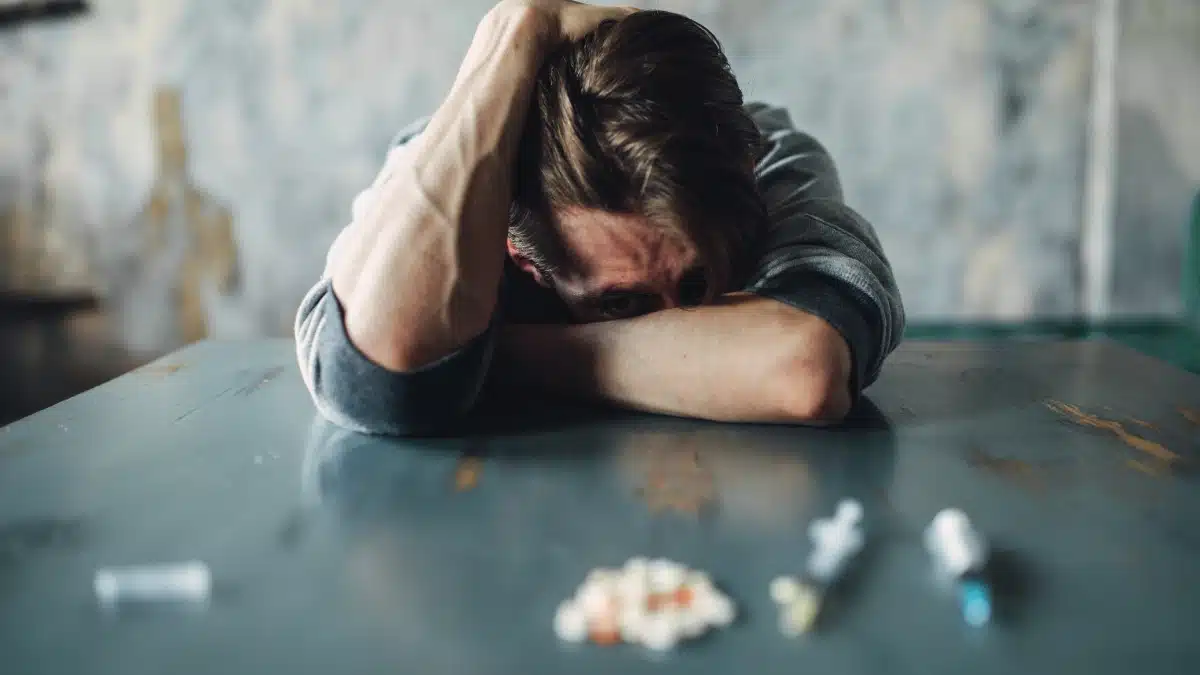 man in dissaray with his head down in his arms and with various drugs in front of him on a table