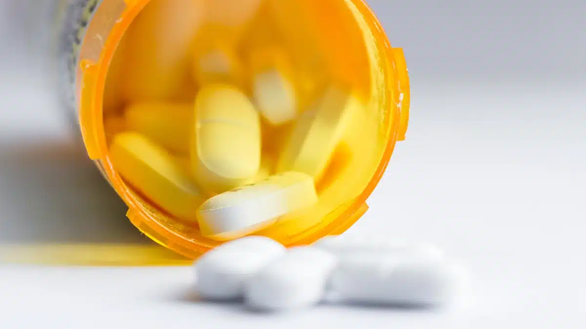 A close-up of a prescription pill bottle tipped over, with yellow and white tablets spilling out onto a surface