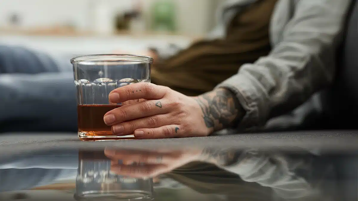 A close-up of a person's hand holding a glass of alcohol while sitting, with their tattooed hand resting on a table that reflects the glass