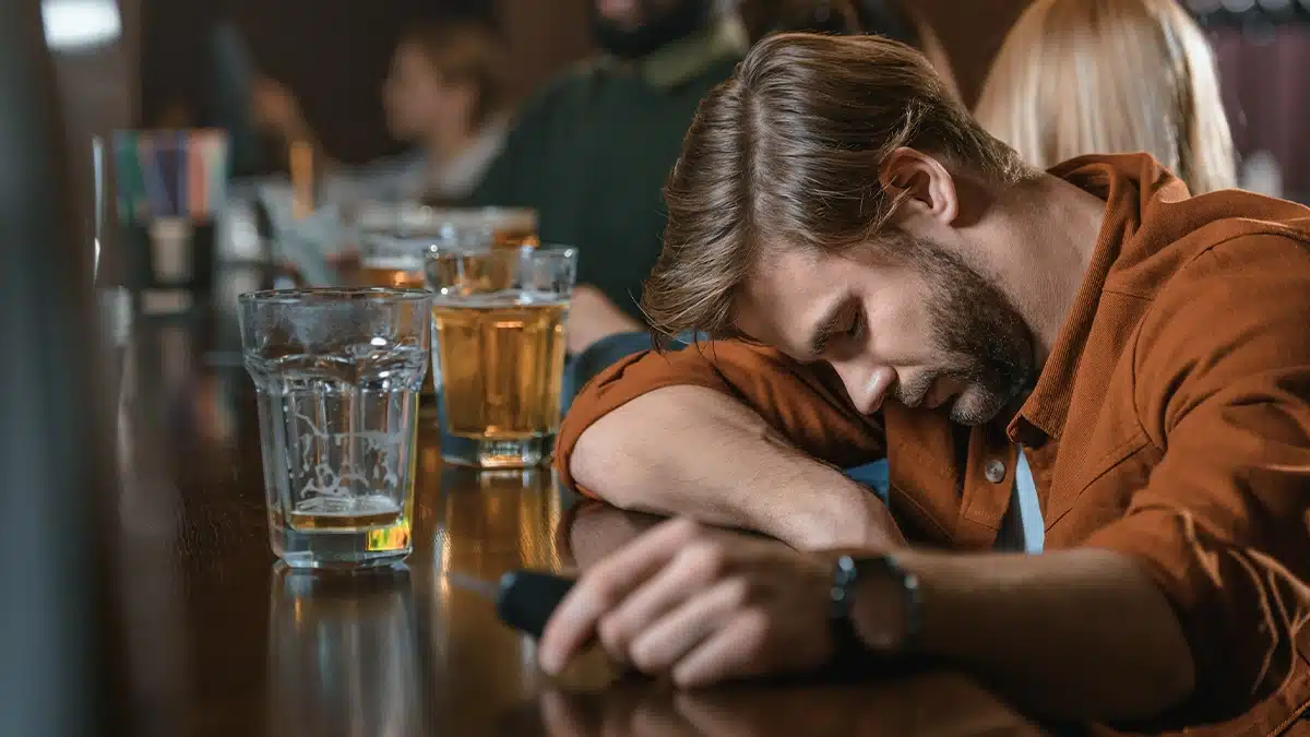 A man with his head down on a bar counter appears to be asleep or passed out. Several empty glasses of alcohol are lined up in front of him. The bar is dimly lit, and there are other patrons in the background, but the focus is on the man in the foreground, suggesting a sense of exhaustion or the effects of heavy drinking.