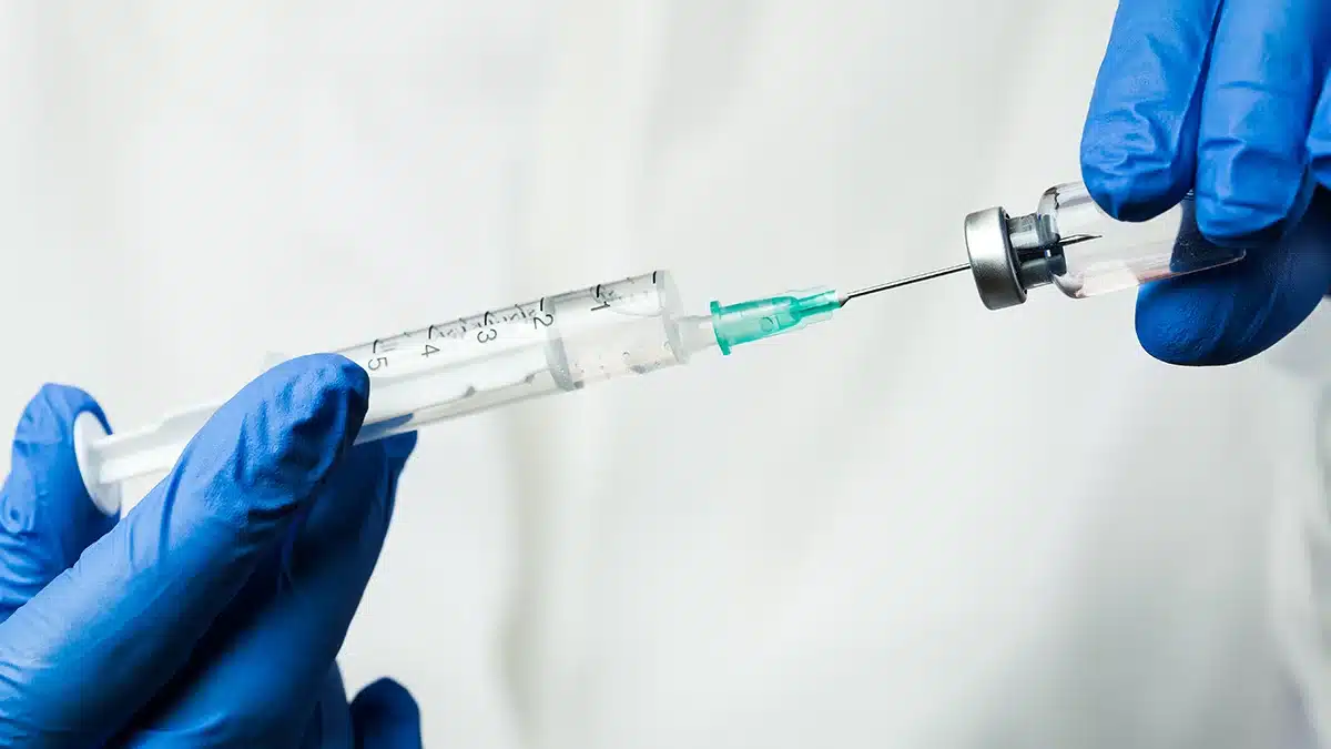 A close-up image of hands wearing blue gloves holding a syringe and a vial. The syringe is being filled with liquid from the vial against a neutral background