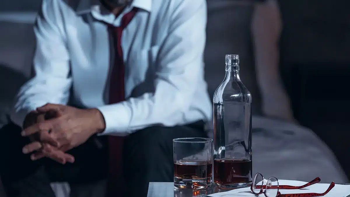 A man dressed in a white shirt and red tie sits with his hands clasped in front of him, looking down. In the foreground, there is an almost empty bottle of alcohol, a glass with some alcohol, and a pair of glasses on a table, suggesting a somber or reflective mood