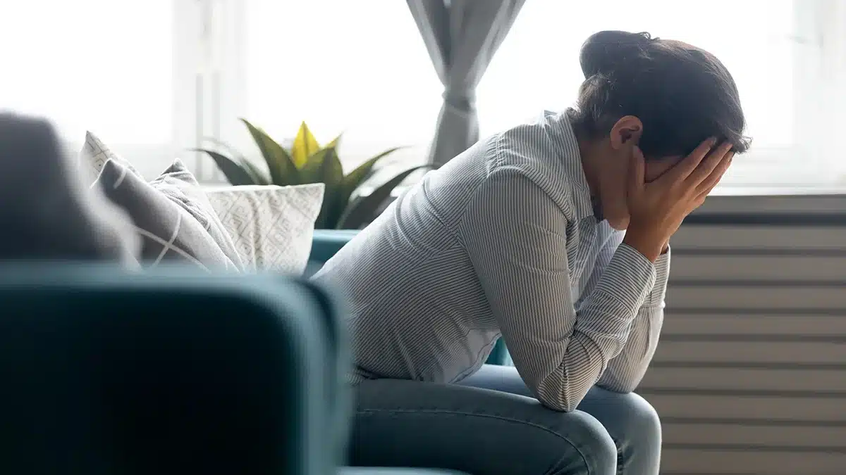 A woman sitting on a couch, leaning forward with her head in her hands, appearing stressed or overwhelmed, in a bright, softly lit room