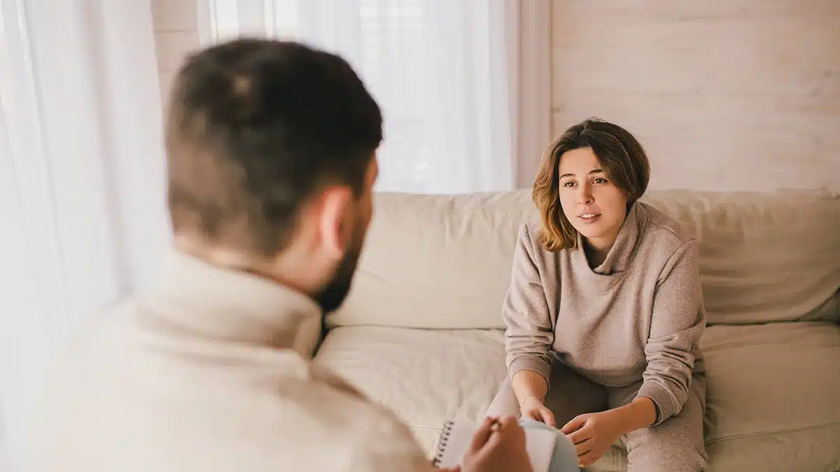 A woman sits on a couch during a therapy session, speaking to a therapist who is taking notes, focusing on her facial expression as she engages in the conversation