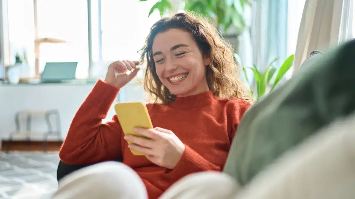 A woman sitting on a couch, smiling and looking at her smartphone, appearing happy and engaged in a conversation or activity on her phone