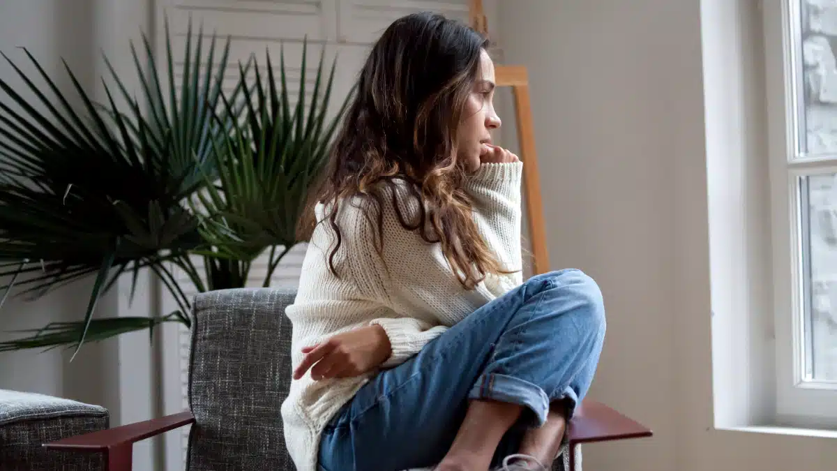 A young woman sitting in a chair, curled up with her knees close to her chest, gazing out of a window with a contemplative expression, suggesting a moment of introspection or emotional difficulty