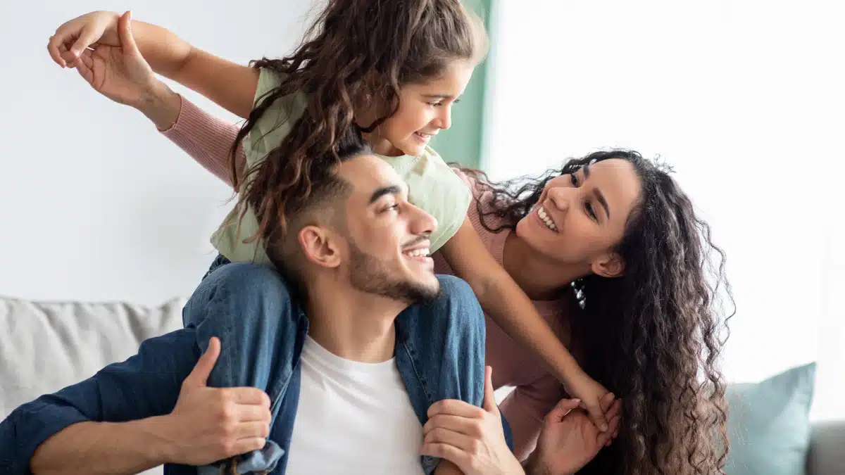 A happy family at home, with a father giving his young daughter a piggyback ride while the mother smiles and interacts with them, creating a warm and joyful moment.