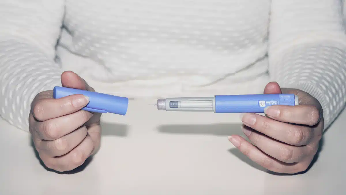 A person holding an insulin pen with a blue casing, partially uncapped to reveal the needle, preparing to administer an injection