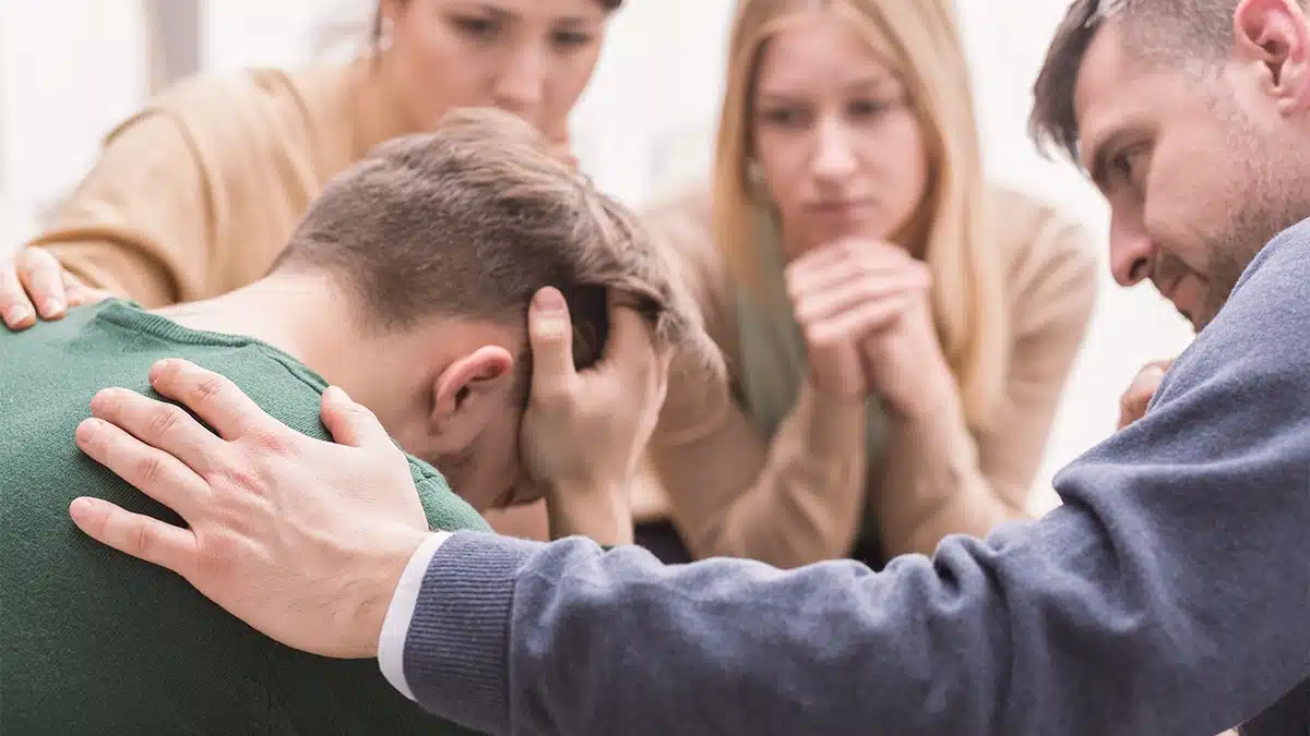 A group of people is gathered closely around a distressed individual who is sitting with his head in his hands.