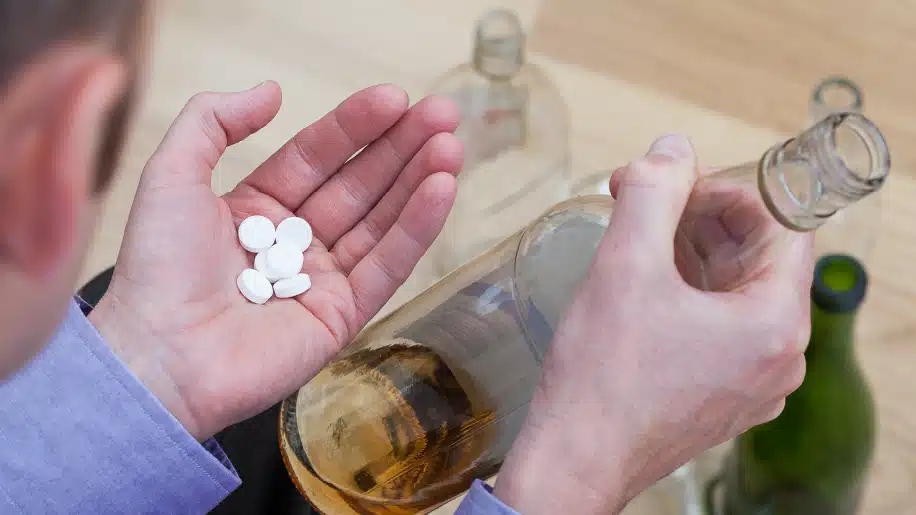 The image shows a person holding a bottle of alcohol in one hand and several white pills in the other, highlighting the dangerous combination of alcohol and prescription medication or illicit drugs.