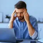 A man sitting at a desk in front of a laptop, appearing stressed or deep in thought.