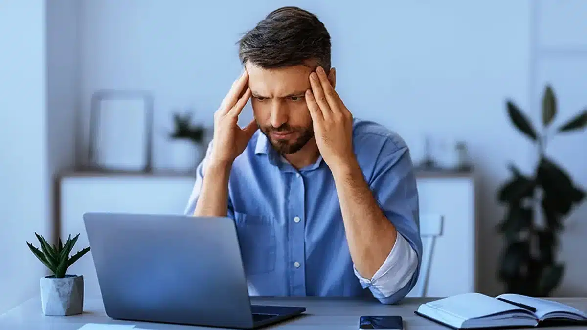 A man sitting at a desk in front of a laptop, appearing stressed or deep in thought.