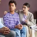 The image shows a teenage boy sitting with two adults in what appears to be a counseling or therapy session. The boy is wearing a purple and white striped long-sleeve shirt and blue jeans, looking slightly concerned or contemplative. One of the adults, a woman sitting beside him, has her arm around his shoulders and is speaking, possibly offering support or advice. The other adult, likely a counselor or therapist, is partially visible, holding a pen and notebook, and seems to be listening attentively. The setting is a calm, indoor environment, likely an office or therapy room.