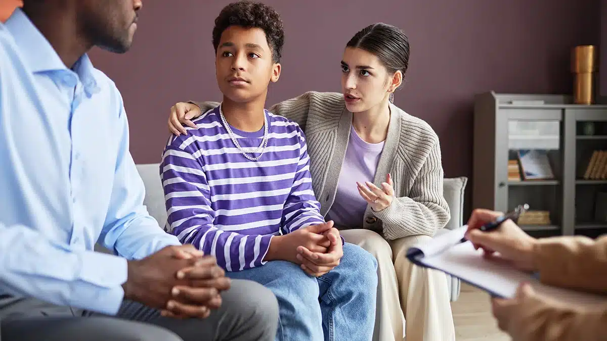 The image shows a teenage boy sitting with two adults in what appears to be a counseling or therapy session. The boy is wearing a purple and white striped long-sleeve shirt and blue jeans, looking slightly concerned or contemplative. One of the adults, a woman sitting beside him, has her arm around his shoulders and is speaking, possibly offering support or advice. The other adult, likely a counselor or therapist, is partially visible, holding a pen and notebook, and seems to be listening attentively. The setting is a calm, indoor environment, likely an office or therapy room.