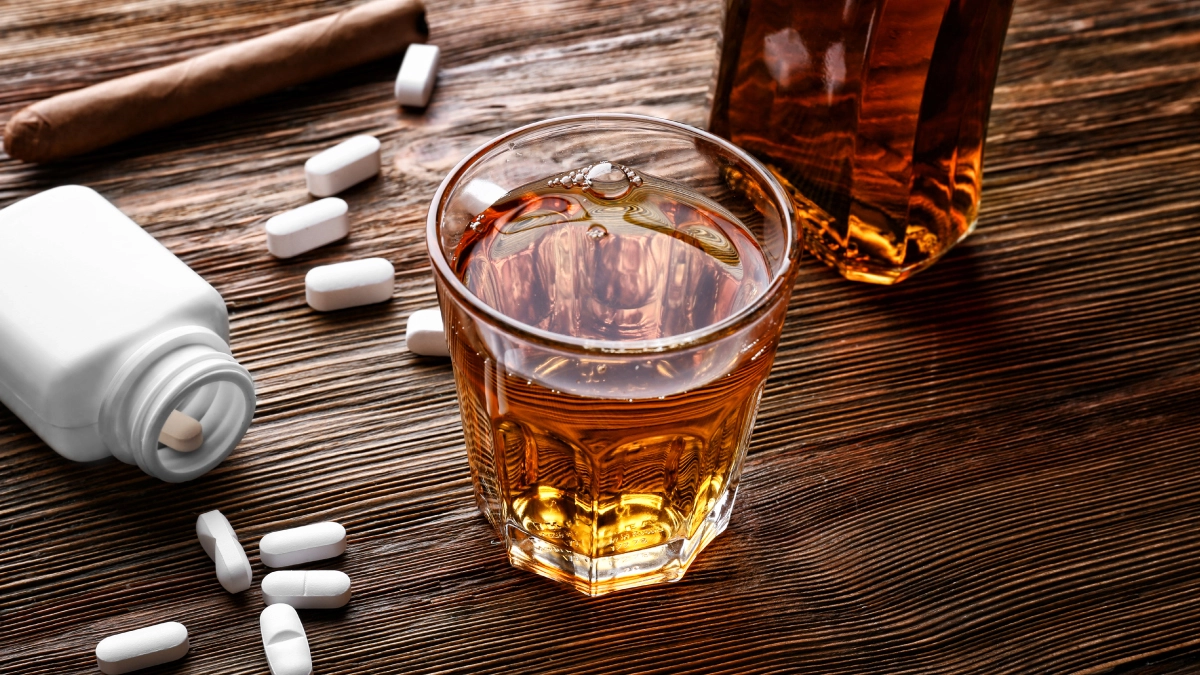 a close-up of a glass of whiskey placed on a wooden surface, with a bottle of alcohol partially visible beside it. Scattered around the glass are several white pills and a white pill bottle lying on its side