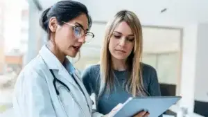 A female doctor in a white coat and stethoscope is reviewing information on a tablet with a female patient. They appear to be in a bright, modern medical office, both focused and engaged in the discussion.
