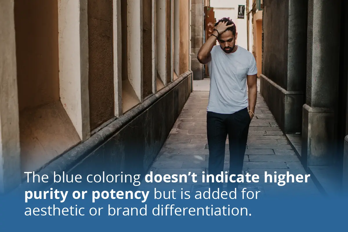 A man in a white t-shirt and dark pants walks alone down a narrow alley, looking down with one hand on his head, appearing worried or deep in thought. At the bottom of the image, over a blue gradient, white text reads: "The blue coloring doesn’t indicate higher purity or potency but is added for aesthetic or brand differentiation."