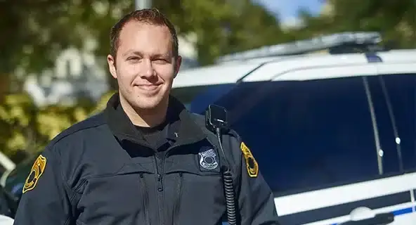 A smiling police officer in a dark uniform stands in front of a patrol car.