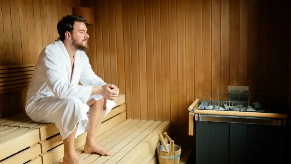 A man in a white bathrobe is sitting on a wooden bench inside a sauna.
