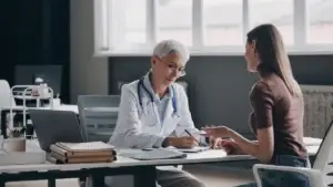 An older female doctor with short gray hair and glasses sits at a desk speaking with a younger female patient. The doctor is writing notes while the patient gestures with her hands. The setting is a modern medical office with large windows and books on the desk.