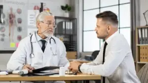 An older male doctor in a white coat with a stethoscope speaks seriously with a male patient in business attire. They are seated across a desk in a medical office setting, with anatomical charts and office supplies visible in the background.