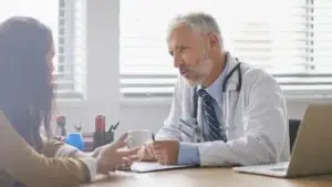 A mature male doctor with gray hair and a stethoscope listens attentively to a female patient seated across from him. They are having a serious conversation at a desk in a well-lit medical office, with papers, a coffee mug, and a laptop nearby.