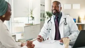 A smiling male doctor in a white lab coat and stethoscope is speaking with a patient across the desk.