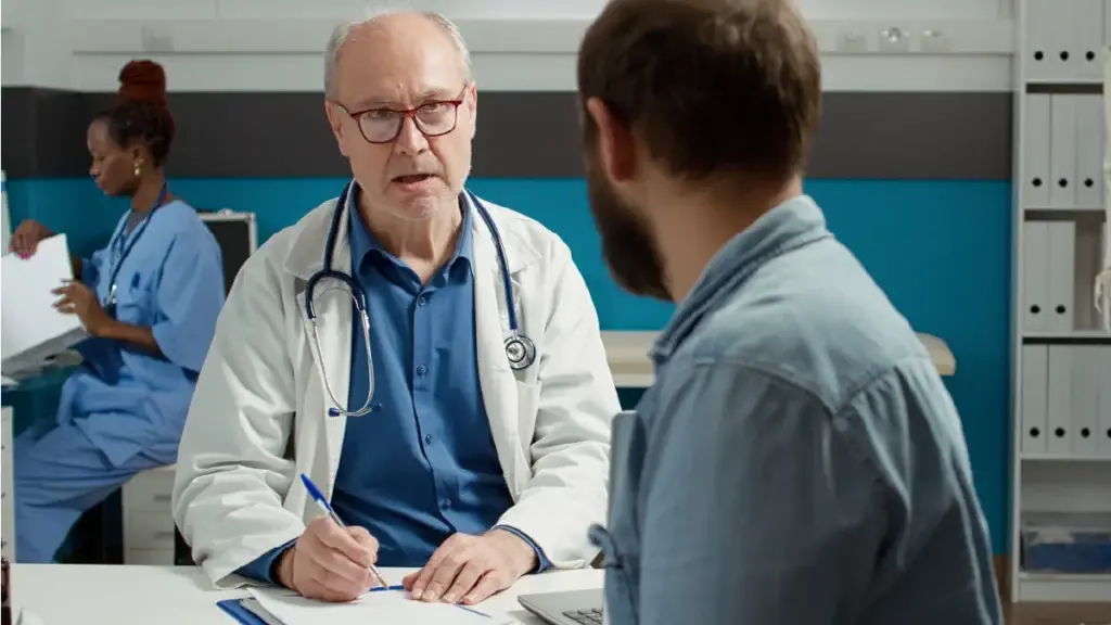 A senior male doctor wearing a white lab coat and stethoscope is having a serious conversation with a younger male patient