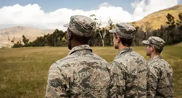 Three military personnel wearing camouflage uniforms and caps stand in an open field, facing away from the camera. They are looking towards a landscape of rolling hills, trees, and a partly cloudy sky.