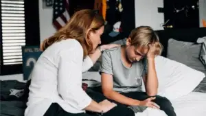 A concerned adult woman sits on a bed next to a distressed teenage boy who is looking down and holding his head. The woman rests a hand on his shoulder, offering comfort in a bedroom setting with sports memorabilia and an American flag in the background.