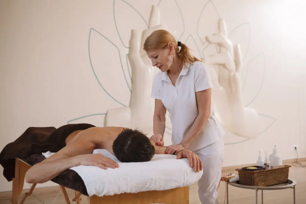 A senior female masseur gives a massage to a male client in a spa setting.