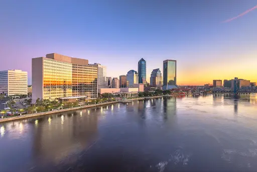 City skyline with modern glass buildings reflected in water at sunset.