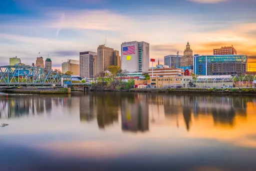 City skyline with tall buildings reflecting in a river at sunset.