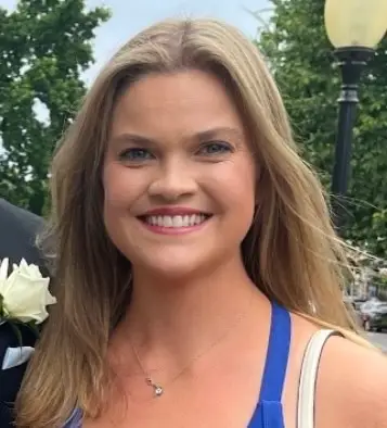 “Smiling woman with long blonde hair wearing a blue dress, standing outdoors with trees and a streetlamp in the background.