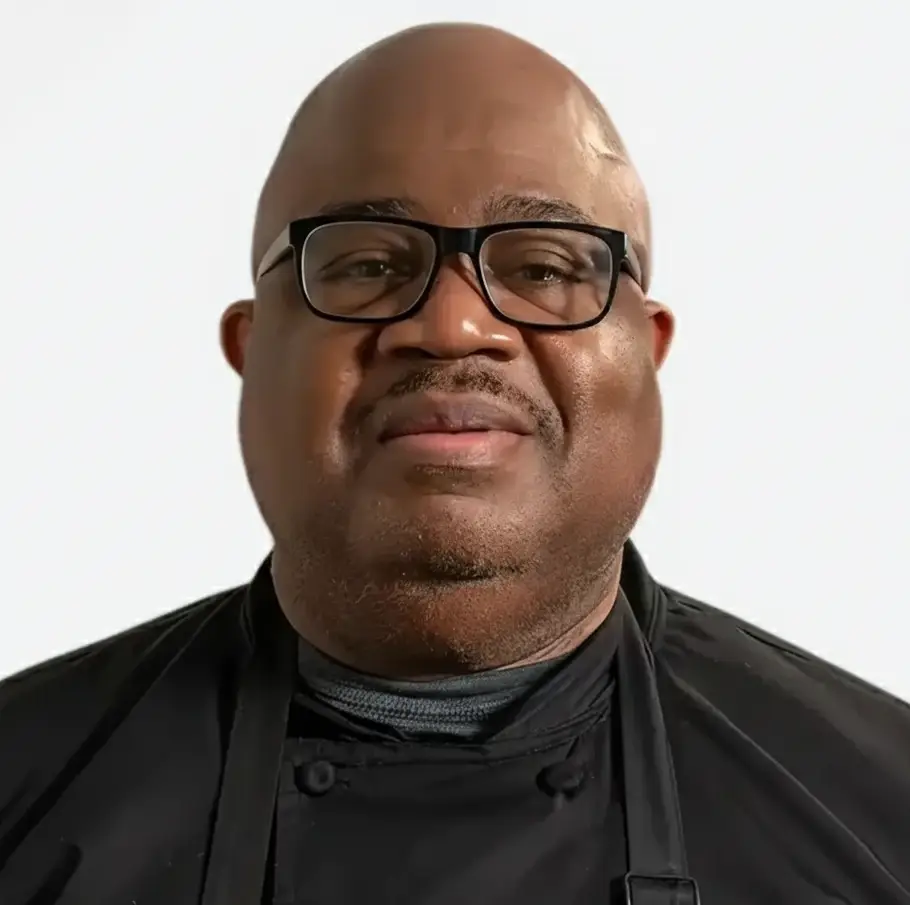 Portrait of a bald man wearing black-framed glasses and a black chef coat with an apron, smiling against a plain light background.