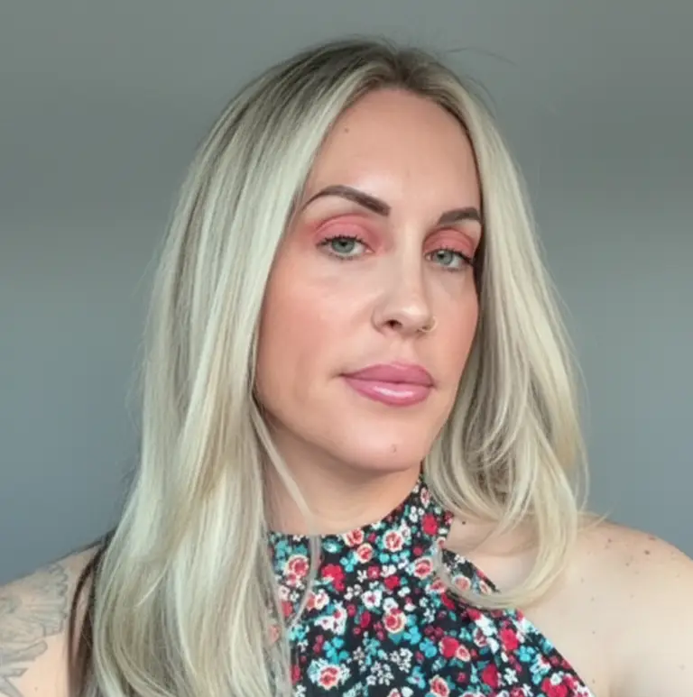 Close-up portrait of a woman with long platinum-blonde hair, pink eye makeup, and glossy lips, wearing a floral halter top against a plain gray background.