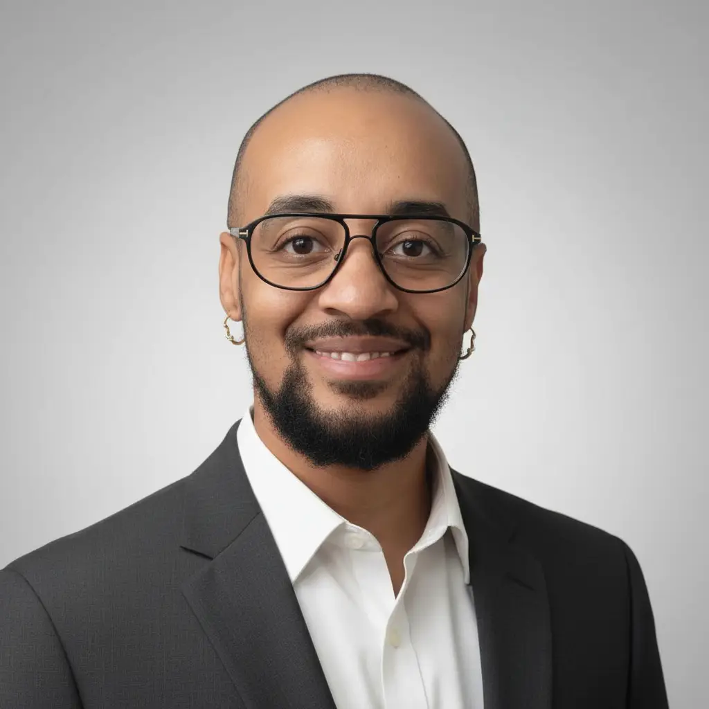 Headshot of a smiling man with a shaved head, black-framed glasses, and small hoop earrings, wearing a dark suit jacket over a white shirt against a light gray background.