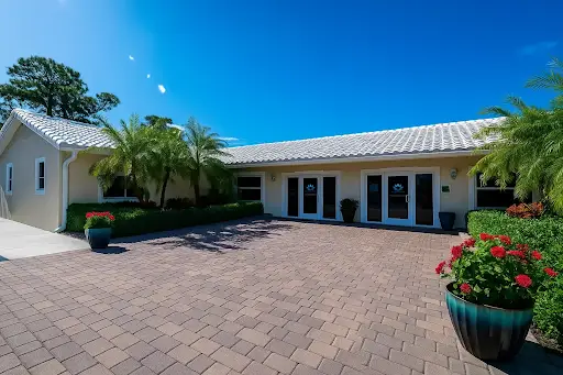Bright, sunny exterior of The Haven Detox Florida with white building, tiled roof, palm trees, and brick-paver courtyard with red flower planters