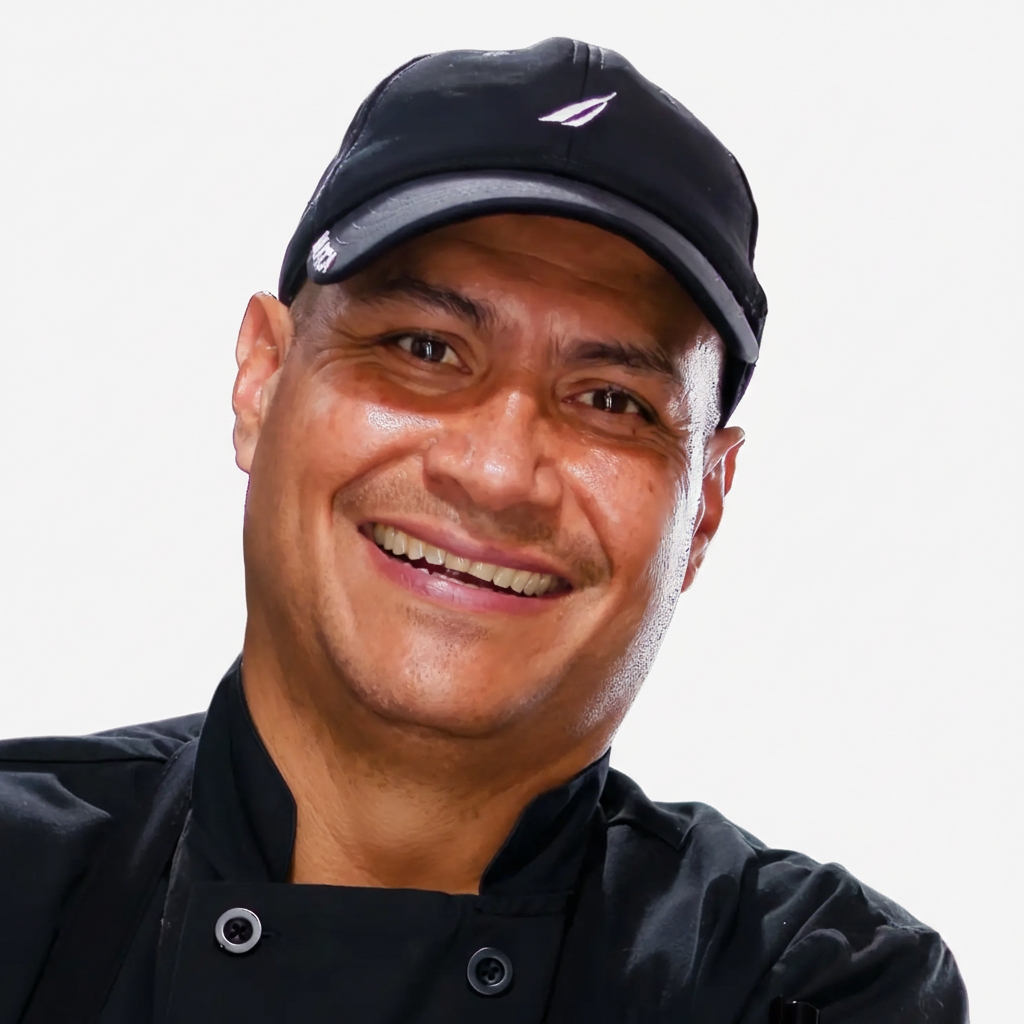 Smiling professional chef wearing a black chef coat and black cap against a clean white background.